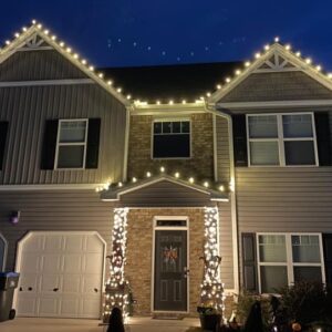 Gray craftsman home with Christmas lights outlining multiple gables and lighted entry shrubs at twilight