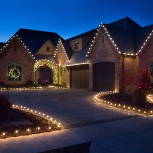 Large brick estate with roofline Christmas lights, lighted wreaths, and a driveway lined with warm white bulbs