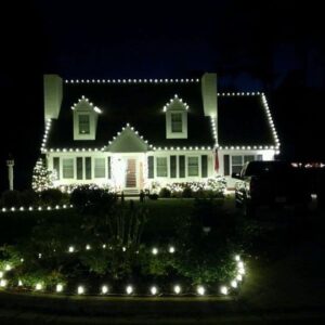 Cape Cod style home with white Christmas lights on the roofline, dormers, and landscape beds